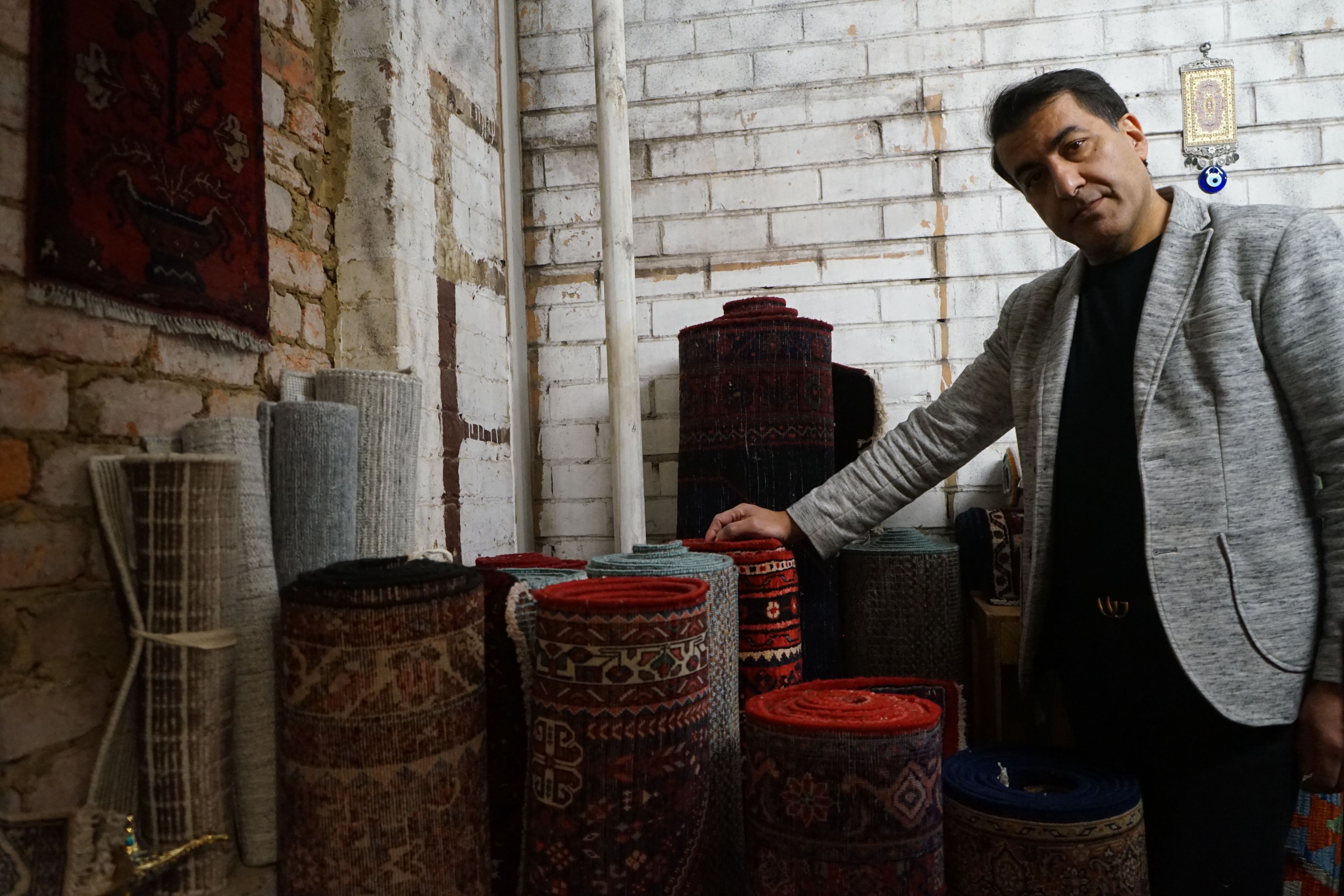 Omid Master in his storeroom, surrounded by rolled up Persian carpets stored vertically, against a white brick wall.