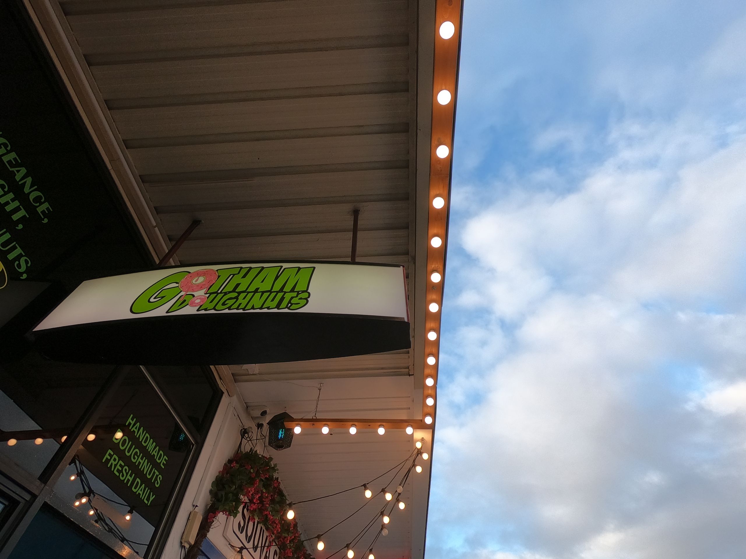 An image looking up at the Gotham Doughnuts street sign. Decorative lights hang along the roof that extends over the footpath. The sky is blue with a few clouds.