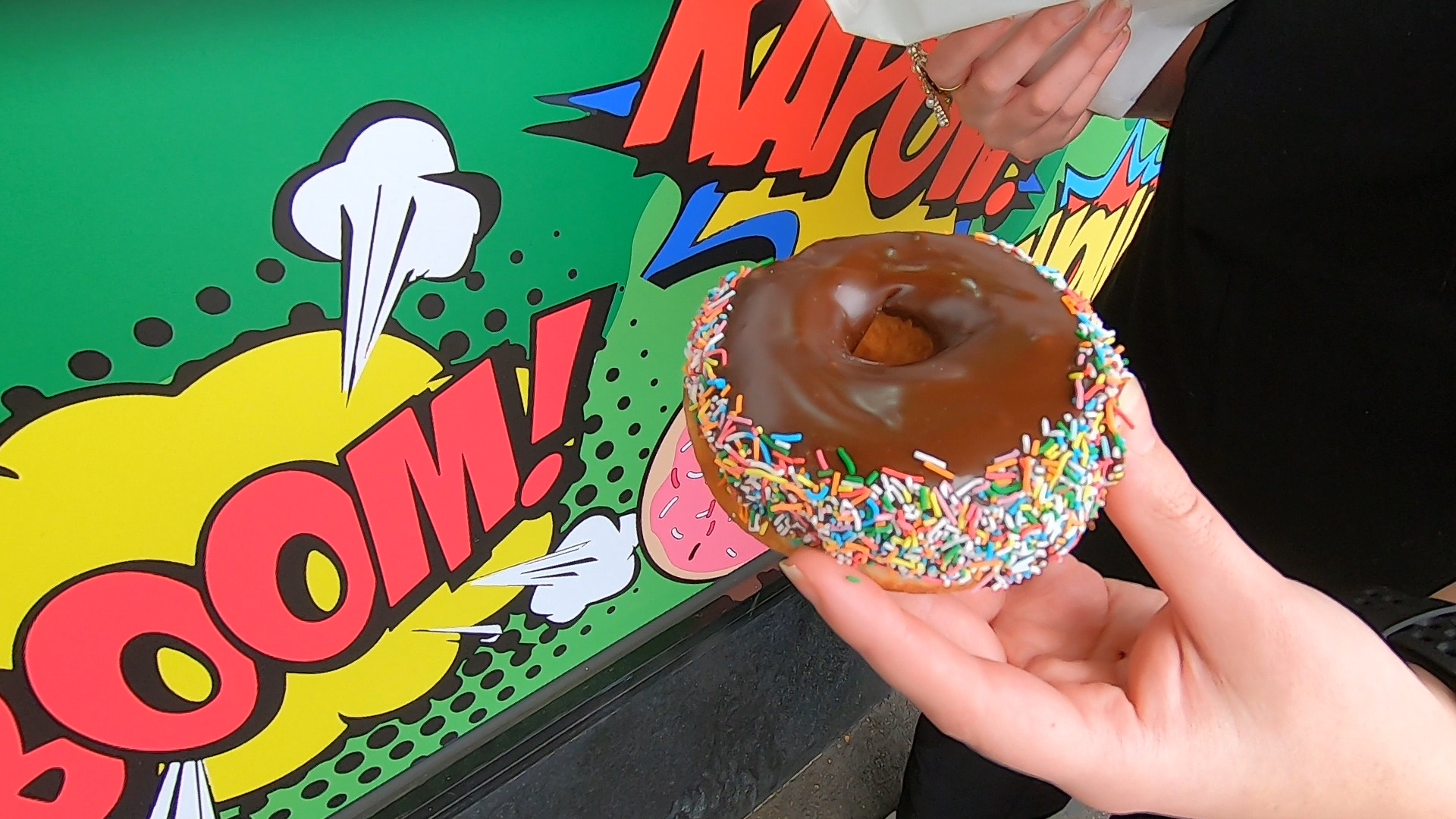 A hand holding a chocolate-iced ring doughnut with sprinkles in front of Gotham Doughnut's graffiti/superhero cartoon style exterior.